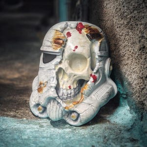 Puede incluir: Un casco de Stormtrooper blanco y desgastado con una calavera reemplazando la cara. El casco tiene detalles color óxido y salpicaduras rojas, lo que sugiere una estética desgastada por la batalla. El diseño combina ciencia ficción con elementos macabros.