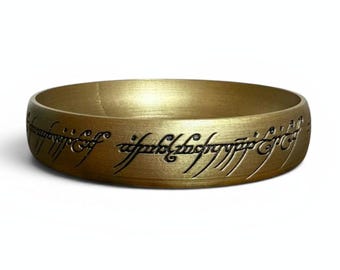 May include: A gold-colored ring with engraved script around the band. The ring has a smooth, metallic finish and is likely a replica or inspired by a fantasy theme.
