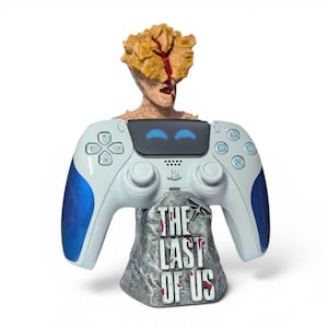 Puede incluir: Un mando de PlayStation 5 blanco y azul con un soporte que muestra "THE LAST OF US". El soporte tiene una apariencia de piedra gris. Una figura que se asemeja a un Clicker del juego está colocada encima del mando.