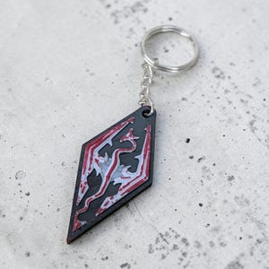 Könnte beinhalten: Schwarzer und roter rautenförmiger Schlüsselanhänger mit einem weißen Drachendesign. Der Schlüsselanhänger hat einen silbernen Schlüsselring und eine Kette. Das Design ist ein stilisierter Drache, ein beliebtes Symbol in Fantasy-Spielen. Der Schlüsselanhänger ist wahrscheinlich aus Holz oder einem ähnlichen Material.