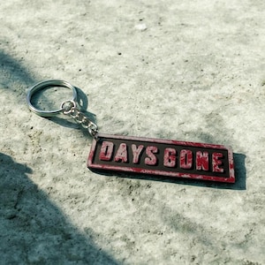 Könnte beinhalten: Ein rechteckiger Schlüsselanhänger mit den Worten "DAYS GONE" in roten Buchstaben auf schwarzem Hintergrund. Der Schlüsselanhänger hat einen silbernen Metallring und eine Kette. Der Hintergrund ist eine strukturierte, hellgraue Oberfläche.