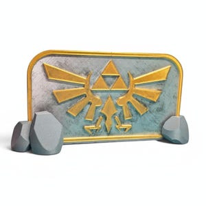 Puede incluir: Un emblema decorativo con el símbolo de la Trifuerza de The Legend of Zelda. El emblema tiene un diseño de Trifuerza dorado sobre un fondo rectangular azul grisáceo con un borde dorado. Dos acentos grises en forma de roca están en el lado izquierdo.