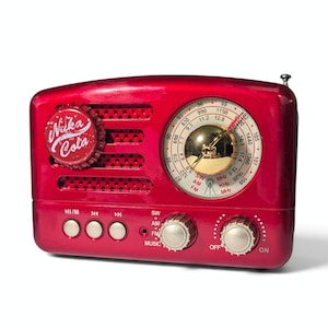 Könnte beinhalten: Ein metallisch rotes tragbares Radio mit einem Nuka Cola-Flaschenverschluss-Design auf dem linken Lautsprechergitter. Die Vorderseite enthält eine runde analoge Skala mit goldener Mitte, drei Tasten, zwei Drehregler und aufgedruckte Beschriftungen.