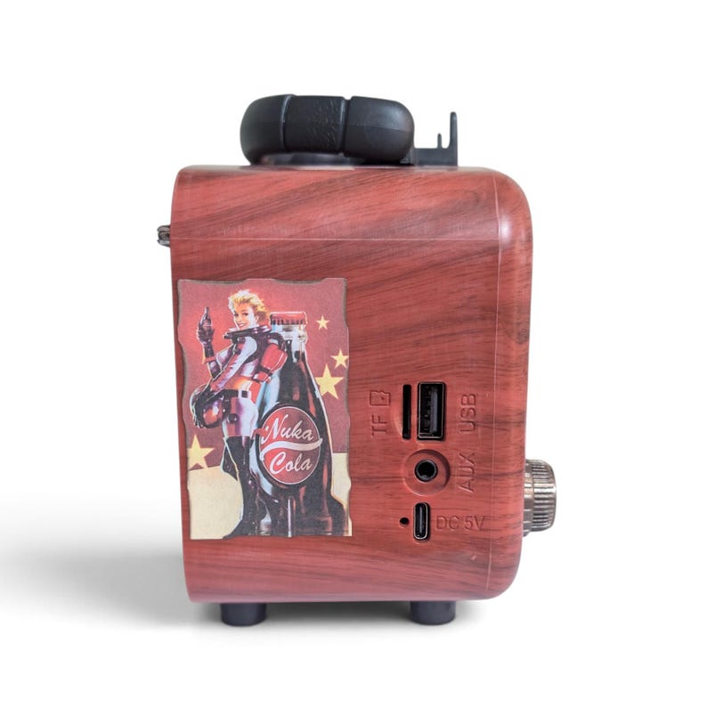 May include: A portable speaker with a faux wood grain finish featuring a Nuka Cola graphic with a character and stars. The side panel includes a TF card slot, a USB port, an AUX input, and a DC 5V charging port. A black handle sits on top.