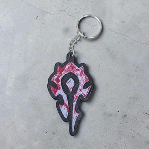 Könnte beinhalten: Ein schwarz-roter Schlüsselanhänger mit dem Horde-Symbol aus dem Videospiel World of Warcraft. Der Schlüsselanhänger hat eine silberne Kette und einen Ring zur Befestigung an Schlüsseln oder einer Tasche. Das Symbol ist ein stilisiertes Design mit schwarzem Umriss und roten und weißen Details.
