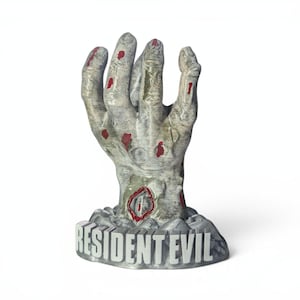 May include: A gray, zombie-like hand statue with red blood-like splatters. The base of the statue reads "RESIDENT EVIL" in gray letters. The hand is reaching upwards, with a detailed, textured appearance.