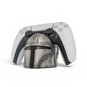 May include: A silver-colored Mandalorian helmet-shaped controller stand cradles a white and black PlayStation 5 controller. The stand is designed to hold the controller securely, showcasing a Star Wars theme.