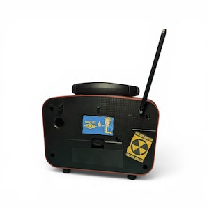 May include: A black and red rectangular radio-shaped device with a single antenna extending upward. The front panel features a Vault-Tec sticker with a cartoon character giving a thumbs up and a yellow Fallout Shelter sticker with a radiation symbol.