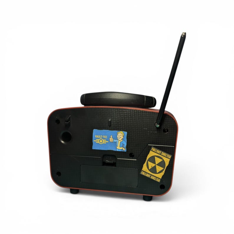 May include: A black and red rectangular radio-shaped device with a single antenna extending upward. The front panel features a Vault-Tec sticker with a cartoon character giving a thumbs up and a yellow Fallout Shelter sticker with a radiation symbol.