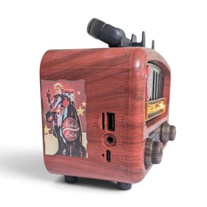 May include: A portable speaker with a faux wood grain finish featuring a Nuka Cola graphic on the side. The device includes a TF card slot, USB port, AUX input, and DC 5V charging port. It has a black handle on top and two circular control knobs.