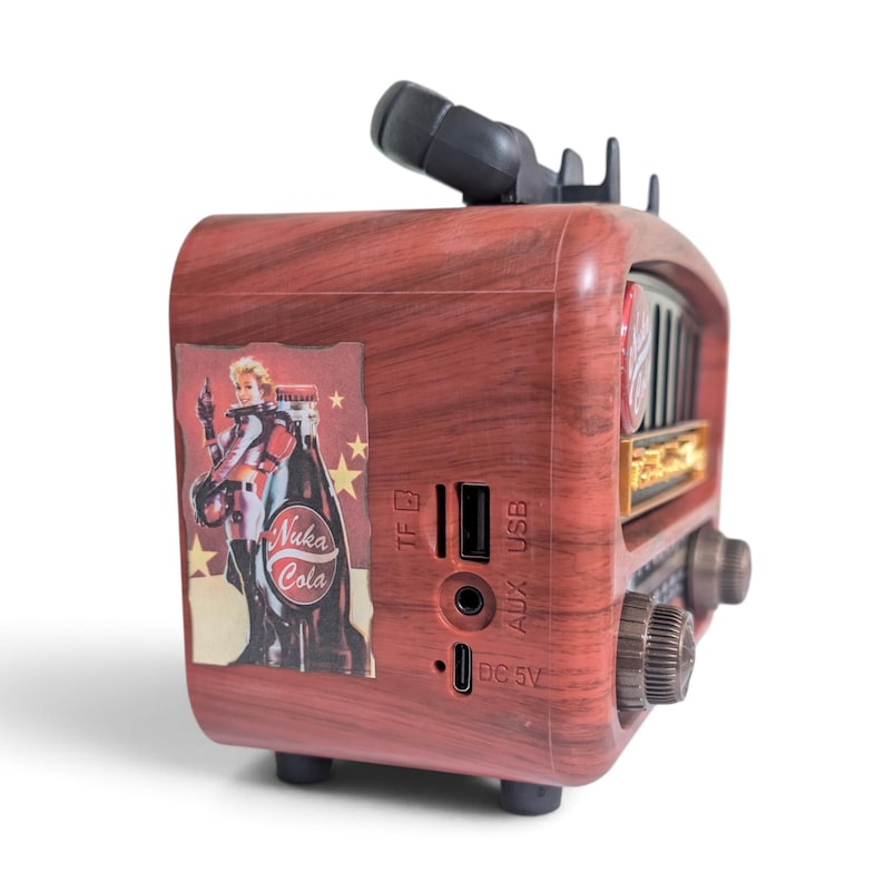 May include: A portable speaker with a faux wood grain finish featuring a Nuka Cola graphic on the side. The device includes a TF card slot, USB port, AUX input, and DC 5V charging port. It has a black handle on top and two circular control knobs.