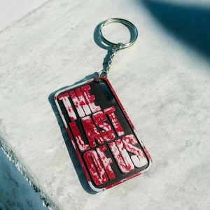 Könnte beinhalten: Ein rechteckiger Schlüsselanhänger mit dem Text "THE LAST OF US" in weißer und roter Schrift. Der Schlüsselanhänger hat einen schwarzen Hintergrund und einen silbernen Schlüsselring.