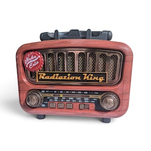 May include: A wood-grain finish radio featuring a Nuka-Cola bottle cap on the top left corner. The front displays the text Radiation King in gold lettering, a speaker grille, two circular control knobs, and a digital frequency display panel.