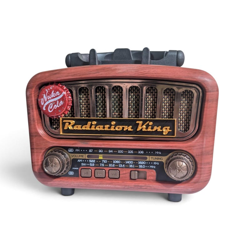 May include: A wood-grain finish radio featuring a Nuka-Cola bottle cap on the top left corner. The front displays the text Radiation King in gold lettering, a speaker grille, two circular control knobs, and a digital frequency display panel.