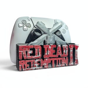 Op de afbeelding: Een witte PlayStation 5-controller met een zilveren embleem en de woorden "RED DEAD REDEMPTION II" in rode en witte letters. De controller staat op de achtergrond, met het embleem en de tekst op de voorgrond.