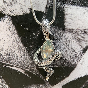 Op de afbeelding: Een zilverkleurige slangenketting met een hanger met een textuur van schubben die om een centrale, gefacetteerde, lichtblauwe ovale edelsteen kronkelt. De hanger hangt aan een dunne, bijpassende zilverkleurige slangenketting met gedetailleerd metaalwerk.