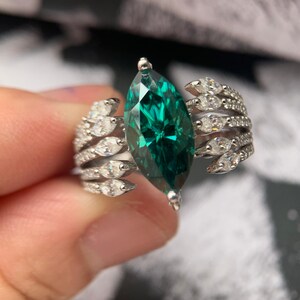 May include: A silver ring with a large, emerald-green marquise-cut gemstone at its centre. The ring's band is decorated with multiple rows of small, clear gemstones, creating a sparkling, elegant design. The ring is held by a person's fingers.