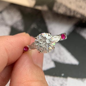 Puede incluir: Un anillo de tono plateado con una gran piedra central redonda y transparente de talla brillante sujeta por cuatro garras. La banda está decorada con dos piedras transparentes más pequeñas de talla marquesa y dos piedras redondas de color rosa intenso.