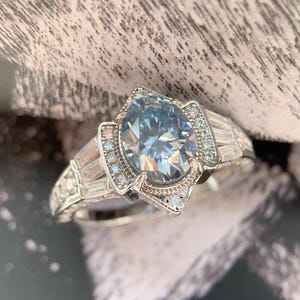 May include: A silver ring featuring a large, light blue, pear-cut gemstone. The ring is embellished with smaller, clear gemstones and has an intricate design. The silver band has a textured pattern. The ring is a piece of jewellery.