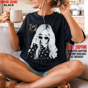 May include: Black Comfort Colors t-shirt featuring a black and white graphic of a woman winking and wearing sunglasses. The shirt has a star pattern on the sleeves and the text "Taylor Swift" in a signature style.