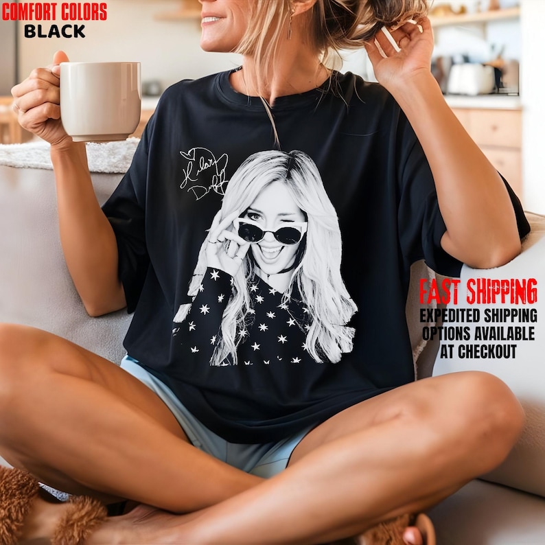 May include: Black Comfort Colors t-shirt featuring a black and white graphic of a woman winking and wearing sunglasses. The shirt has a star pattern on the sleeves and the text "Taylor Swift" in a signature style.