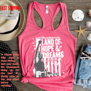 May include: A pink tank top featuring a graphic of a musician with the text "LAND OF HOPE & DREAMS" and "SPRINGSTEEN & E STREET AMERICAN TOUR". The top has a racerback design and is made of a soft material.