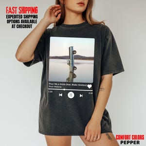 May include: Dark grey t-shirt featuring a rectangular graphic of a truck reflected in water. The graphic includes the text "Pour Me a Drink (feat. Blake Shelton) Post Malone". The shirt has a music player interface design.
