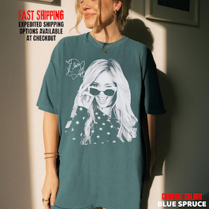 May include: Blue spruce t-shirt featuring a black and white graphic of a woman winking and wearing sunglasses. The shirt also has a signature and star pattern. The text "FAST SHIPPING EXPEDITED SHIPPING OPTIONS AVAILABLE AT CHECKOUT" is visible.
