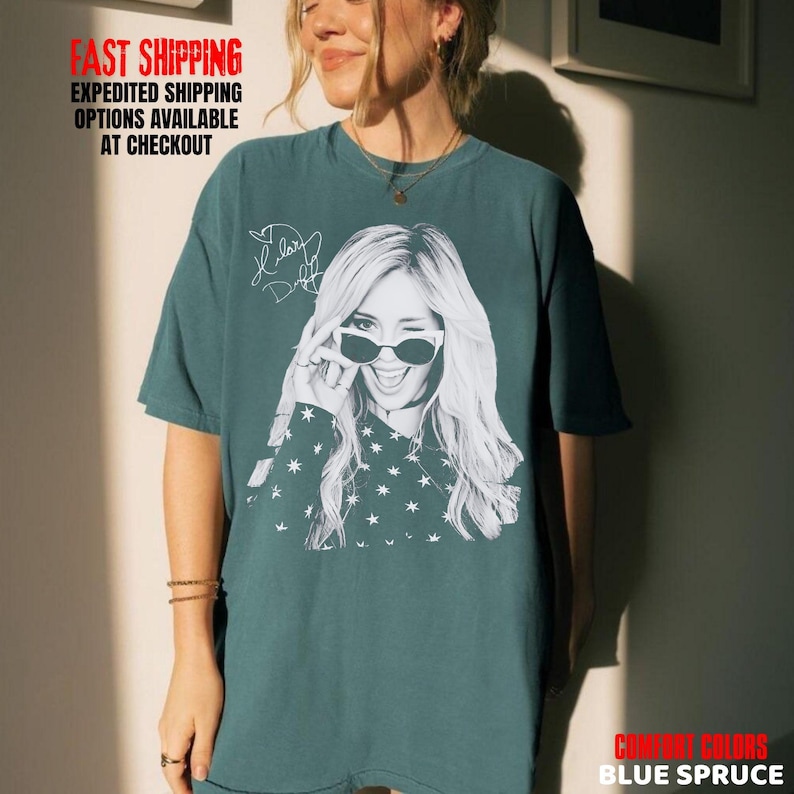 May include: Blue spruce t-shirt featuring a black and white graphic of a woman winking and wearing sunglasses. The shirt also has a signature and star pattern. The text "FAST SHIPPING EXPEDITED SHIPPING OPTIONS AVAILABLE AT CHECKOUT" is visible.