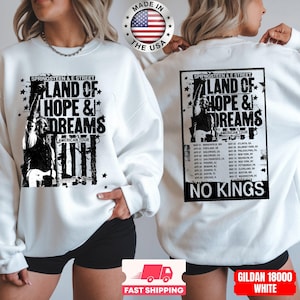 May include: White sweatshirt featuring a black and white graphic with the text "LAND OF HOPE & DREAMS AMERICAN TOUR". The design includes a musician with a guitar and a list of tour dates. The sweatshirt also has the text "NO KINGS".