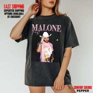 May include: Dark gray t-shirt featuring a graphic of Post Malone wearing a cowboy hat and holding a bottle. The text "MALONE post" is above the image in pink. The shirt is a Comfort Colors Pepper.