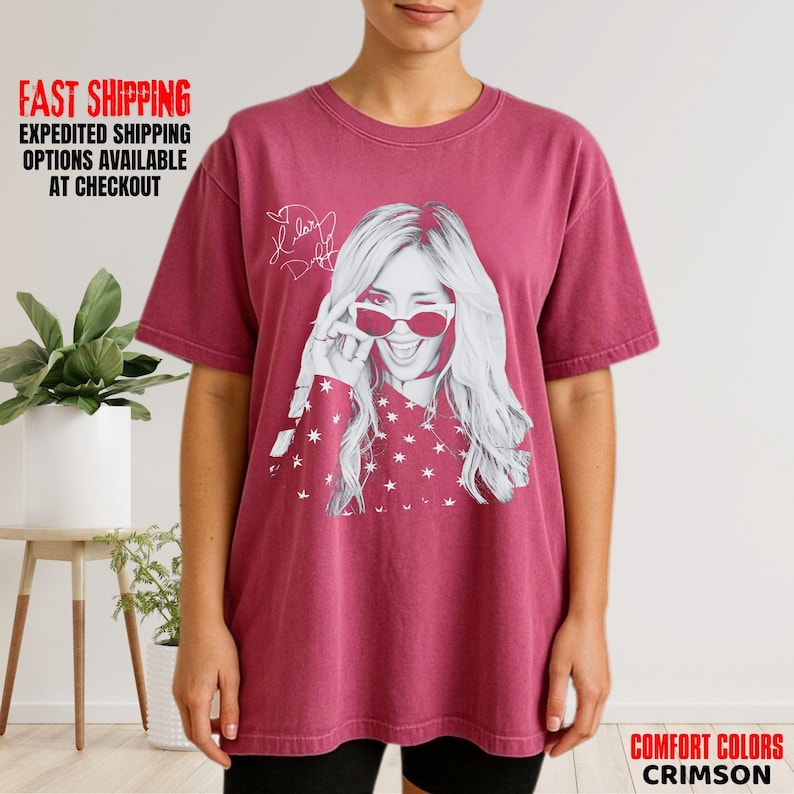 May include: Crimson Comfort Colors t-shirt featuring a black and white image of a woman winking and wearing sunglasses. The image includes the text "Hillary Duff" and a signature. The shirt has a crew neck and short sleeves.