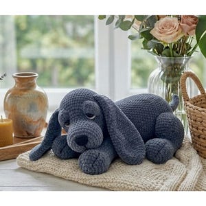 May include: A dark gray crochet dog plush toy with long floppy ears, a rounded snout, and a black nose. The stuffed animal is lying down in a resting position on a cream-colored knitted fabric surface, showcasing a textured yarn construction.