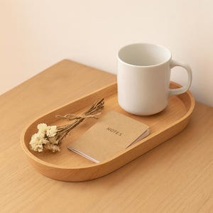 Puede incluir: Una bandeja ovalada de madera clara contiene una taza de cerámica blanca, un pequeño bloc de notas con "NOTES" impreso y flores secas. La bandeja está sobre una superficie de madera clara.