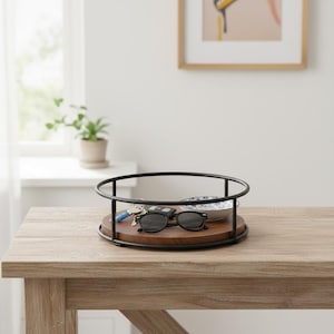 May include: A round, wooden tray with a black metal rim. The tray holds sunglasses, keys, and a small bowl with a blue and white floral pattern. The tray sits on a light brown wooden table.