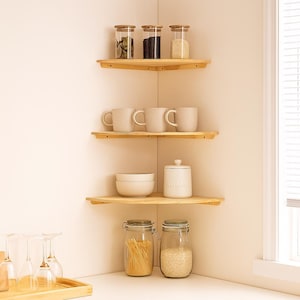 Może przedstawiać: Narożna półka kuchenna z czterema drewnianymi półkami. Półki zawierają szklane słoiki z drewnianymi pokrywkami, kubki, miski i pojemnik na cukier. Słoiki zawierają różne suche produkty, a kubki mają neutralny kolor.