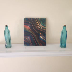 May include: A rectangular canvas with an abstract design in shades of blue, orange, and gold stands between two turquoise glass bottles with cork stoppers. The bottles are rectangular and lean slightly. The items are displayed on a white shelf.