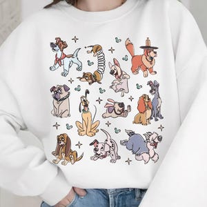 May include: White sweatshirt featuring a colorful illustration of various cartoon dog characters. The dogs are in different poses and colors, with a few stars scattered around the design. The sweatshirt has long sleeves and a crew neck.