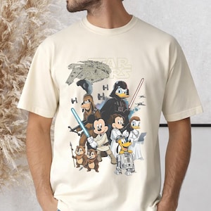 May include: Cream-colored t-shirt featuring a Star Wars graphic with Disney characters. The design includes Darth Vader as Donald Duck, Mickey Mouse as Luke Skywalker, and other characters from both franchises. The Star Wars logo is at the top.