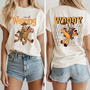 May include: Cream-colored t-shirt featuring a cartoon image of Woody riding a horse, with the text "Sheriff Woody" in orange. The back of the shirt repeats the word "Woody" with the same graphic.