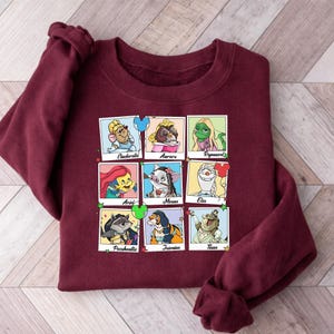 Op de afbeelding: Een bordeauxrode sweatshirt met een raster van negen cartoonkarakterportretten. De karakters zijn onder meer Assepoester, Aurora, Rapunzel, Ariel, Moana, Elsa, Pocahontas, Jasmine en Tiana. Elk portret staat in een vierkant met de naam van het personage eronder.