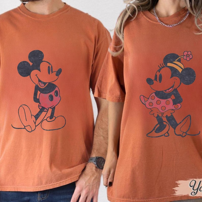 May include: Two orange t-shirts featuring cartoon characters. One shirt displays Mickey Mouse with a red shorts and black outline. The other shirt shows Minnie Mouse in a polka dot skirt and a flower hat. The shirts are likely made of cotton.
