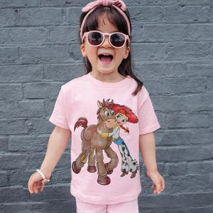 Op de afbeelding: Een roze t-shirt met een cartoonillustratie van een bruin paard en een roodharige cowgirl die elkaar omhelzen. Het kind draagt een roze zonnebril en een roze haarband. Het kind draagt ook een roze short.