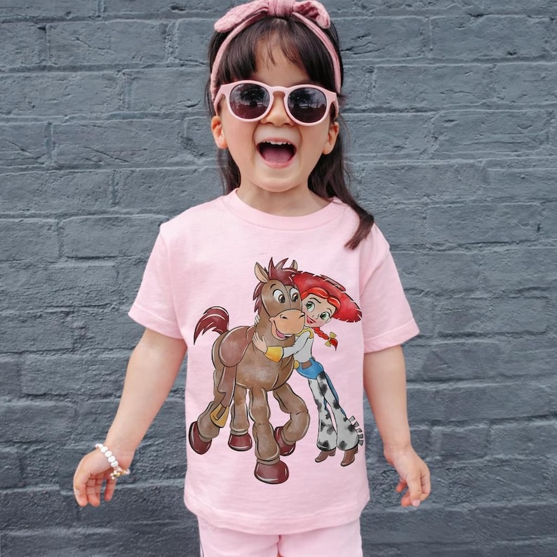 Op de afbeelding: Een roze t-shirt met een cartoonillustratie van een bruin paard en een roodharige cowgirl die elkaar omhelzen. Het kind draagt een roze zonnebril en een roze haarband. Het kind draagt ook een roze short.