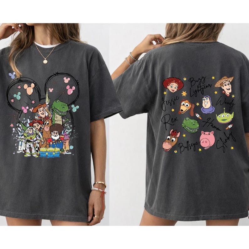 May include: Dark gray t-shirt featuring a Disney-themed design. The front displays a Mickey Mouse silhouette with Toy Story characters. The back showcases individual character portraits with their names, including Buzz Lightyear and Woody.
