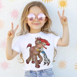 Op de afbeelding: Wit T-shirt met een cartoon illustratie van een bruin paard en een meisje met rood haar. Het meisje omhelst het paard. De afbeelding staat op een wit t-shirt.