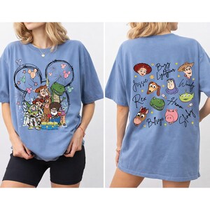 May include: A light blue t-shirt featuring a graphic of Toy Story characters, including Woody, Buzz Lightyear, and Rex, with a Mickey Mouse outline. The back of the shirt displays character names. A comfortable, casual style.