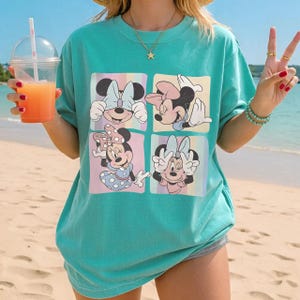 May include: Teal-colored t-shirt featuring a four-panel graphic of Minnie Mouse in various poses. Each panel has a pastel background. The shirt is a loose fit, and the overall style is casual and beachy.
