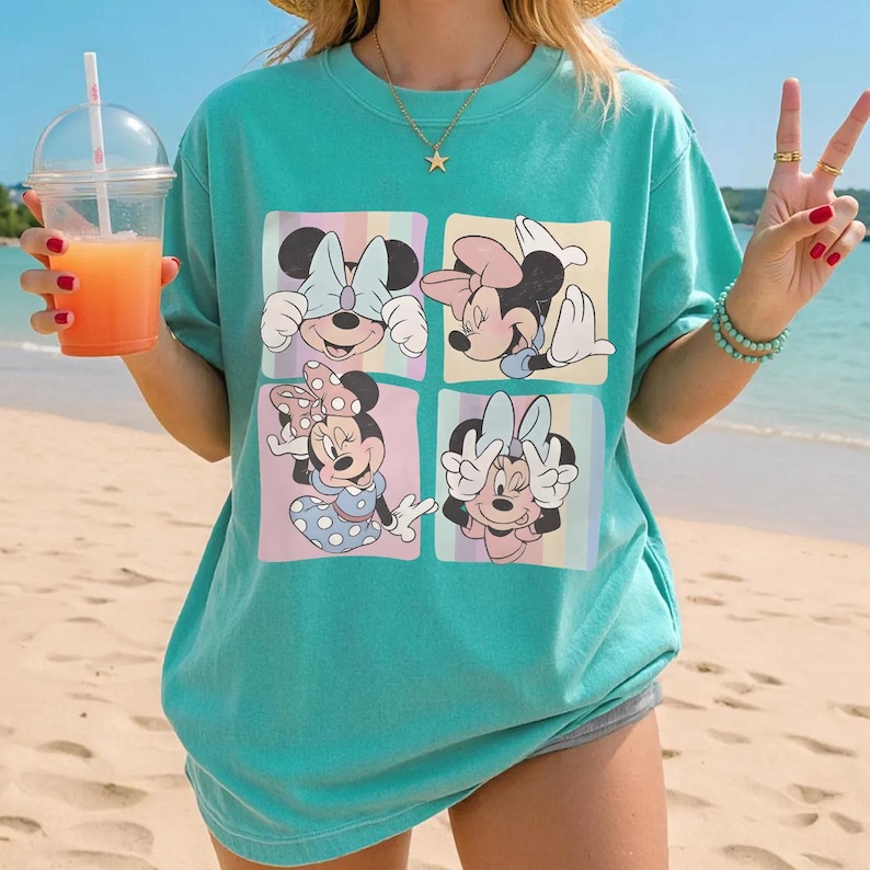 May include: Teal-colored t-shirt featuring a four-panel graphic of Minnie Mouse in various poses. Each panel has a pastel background. The shirt is a loose fit, and the overall style is casual and beachy.