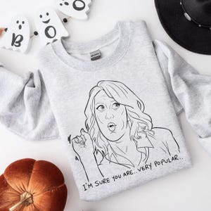 以下が含まれることがあります： 「I'M SURE YOU ARE... VERY POPULAR..」の文字が入った、女性の白黒線画が描かれたライトグレーのスウェットシャツ。スウェットシャツは、カボチャとハロウィーンの装飾が施されています。